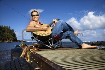 Young Man Sitting Lakeside and Playing Guitar