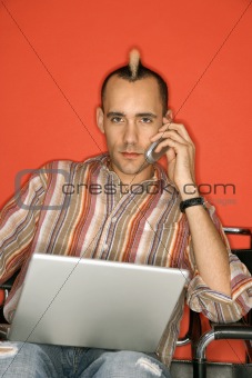 Caucasian man with mohawk using laptop talking on cellphone.