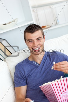 Cheerful young man eating popcorn holding a remote sitting on a sofa in the living room