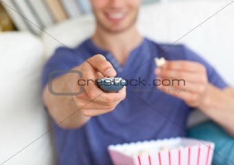 Close-up of a cheerful young man eating popcorn holding a remote sitting on a sofa in the living room