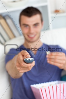 Cheerful young man eating popcorn holding a remote sitting on a sofa in the living room