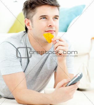 Jolly young man holding a remote eating crisps lying on the fllo