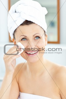 Smiling young woman with a towel putting cream on her face in th