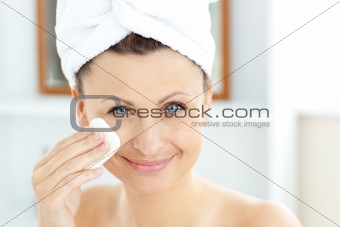 Joyful young woman with a towel putting cream on her face in the