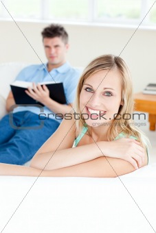 Girlfriend smiling at the camera while her boyfriend is reading 