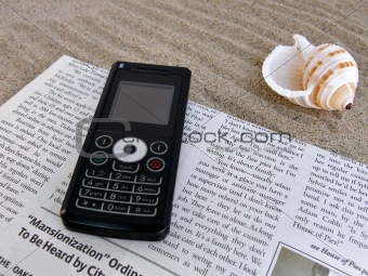 Close up shot of newspaper, celfone, shell on the sand