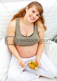 Happy beautiful pregnant woman sitting on sofa with fruit salad  in hand.
