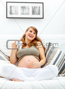 Laughing beautiful pregnant woman relaxing on couch and  holding cup of tea in hand

