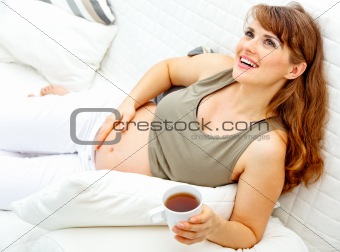 Smiling beautiful pregnant woman relaxing on couch and  holding cup of tea in hand
