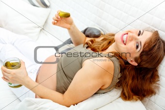 Smiling beautiful pregnant woman relaxing on sofa and  holding jar of pickles in hands
