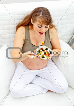 Smiling beautiful pregnant woman sitting on sofa and eating fresh vegetable salad
