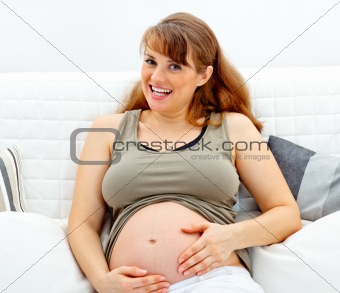 Smiling beautiful pregnant female sitting on sofa and touching her belly
