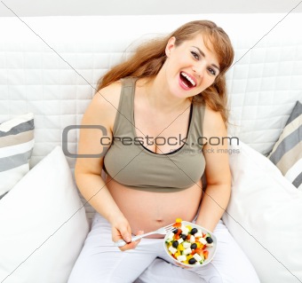 Smiling beautiful pregnant woman sitting on sofa with fresh vegetable salad in hand
