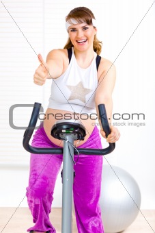 Smiling beautiful pregnant woman working out on static bicycle and showing thumbs up gesture
