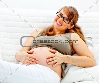 Smiling beautiful pregnant woman sitting on sofa and holding her belly
