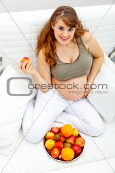 Smiling  beautiful pregnant woman sitting  on sofa and  holding fruit in hand
