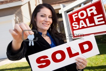 Happy Attractive Hispanic Woman Holding Sold Real Estate Sign and Keys in Front For Sale Real Estate Sign and House.