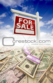 Stacks of Money Fading Off and For Sale Real Estate Sign Against Blue Sky with Clouds.