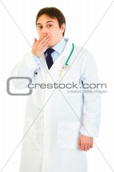Shocked young medical doctor holding hand near mouth 
