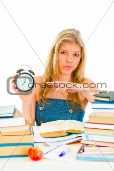 Worried teengirl sitting at table with books and pointing finger on alarm clock
