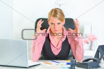 Angry business woman sitting at office desk and holding hands near head
