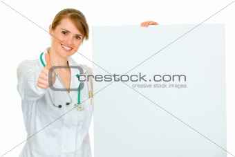 Smiling medical female doctor holding blank billboard and showing thumbs up gesture
