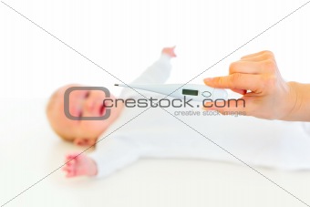 Woman's hand holding thermometer and crying baby laying on back  in background
