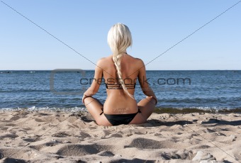 blonde girl in a black bathing suit sits on the beach and meditate