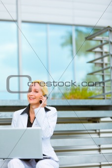 Smiling business woman with laptop sitting on stairs at office building and talking on mobile
