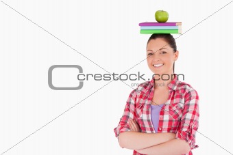 Pretty female holding an apple and books on her head while stand