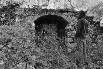 hooded man next to arched recess of rural ruin