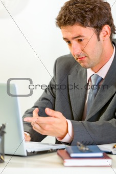 Confused modern businessman sitting at office desk working on laptop
