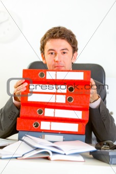 Sitting at office desk confused businessman looking out from pile of folders
