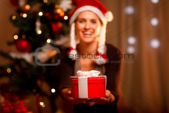 Hand presenting gift box and smiling woman and Christmas tree in background
