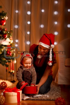Young mother with happy baby opening present box near Christmas tree
