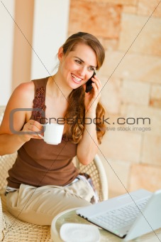 Smiling woman sitting on terrace with laptop and cup of tea and speaking mobile 
