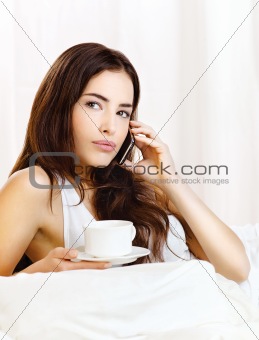 woman making phone call and holding cup of coffee in bed