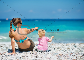 Mother sitting with baby on sea shore and pointing on copy space. Rear view