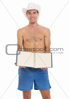 On vacation young man in shorts and beach hat showing blank photo album