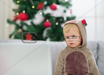Thoughtful baby in deer suit near Christmas tree looking in laptop