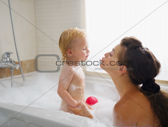 Baby kissing mother while washing in foam filled bathtub