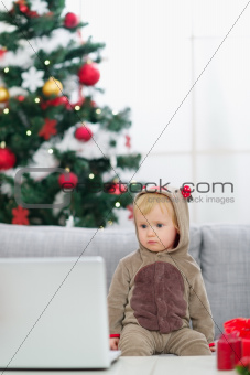 Baby in Christmas deer costume near Christmas tree looking in laptop