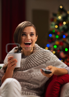 Surprised young woman with cup of hot beverage looking TV