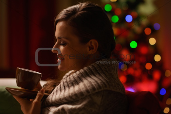 Happy woman sitting chair and drinking hot beverage in front of Christmas tree