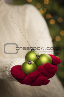 Woman Wearing A Sweater and Seasonal Red Mittens Holding Three Green Christmas Ornaments.
