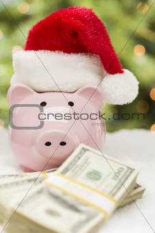Pink Piggy Bank Wearing Santa Hat Near Stacks of Hundreds of Dollars of Money on Snowflakes.