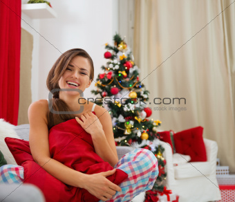 Smiling young woman in pajamas sitting on sofa near Christmas tree