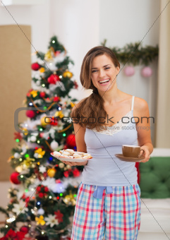 Happy young woman in pajamas with Christmas snacks near Christmas tree