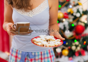 Closeup on woman in pajamas holding hot tea and cookies in front of Christmas tree