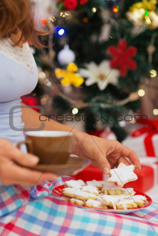 Closeup on woman in pajamas holding hot chocolate and cookies in front of Christmas tree